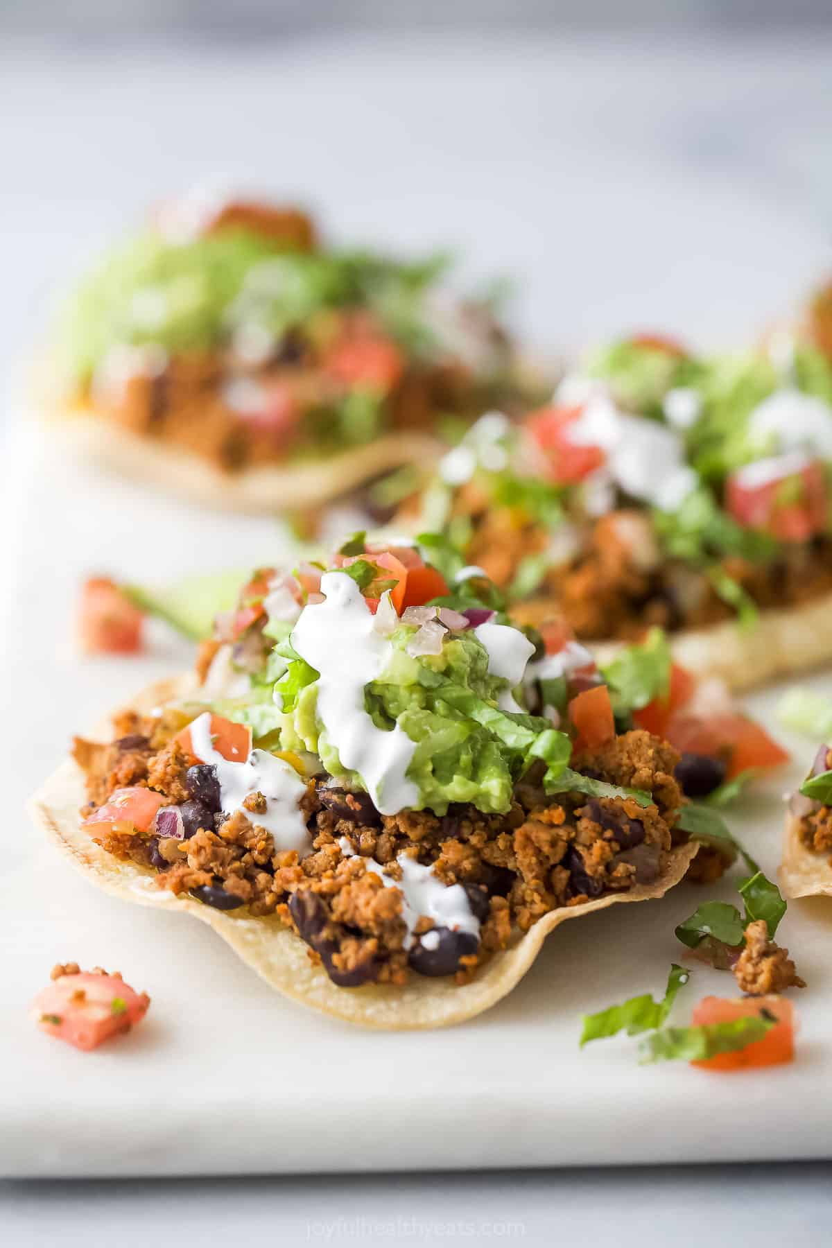 This turkey-black bean tostadas recipe with smokey Tex-Mex seasonings and fresh veggie toppings is the perfect weeknight dinner. Assembled turkey tostadas recipe with sour cream, diced tomatoes, shredded lettuce, and guacamole on top.