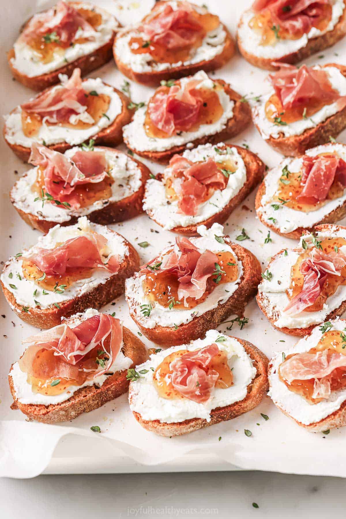 Angled photo of crunchy crostinis with creamy goat cheese, fig jam, and salty prosciutto on top. 