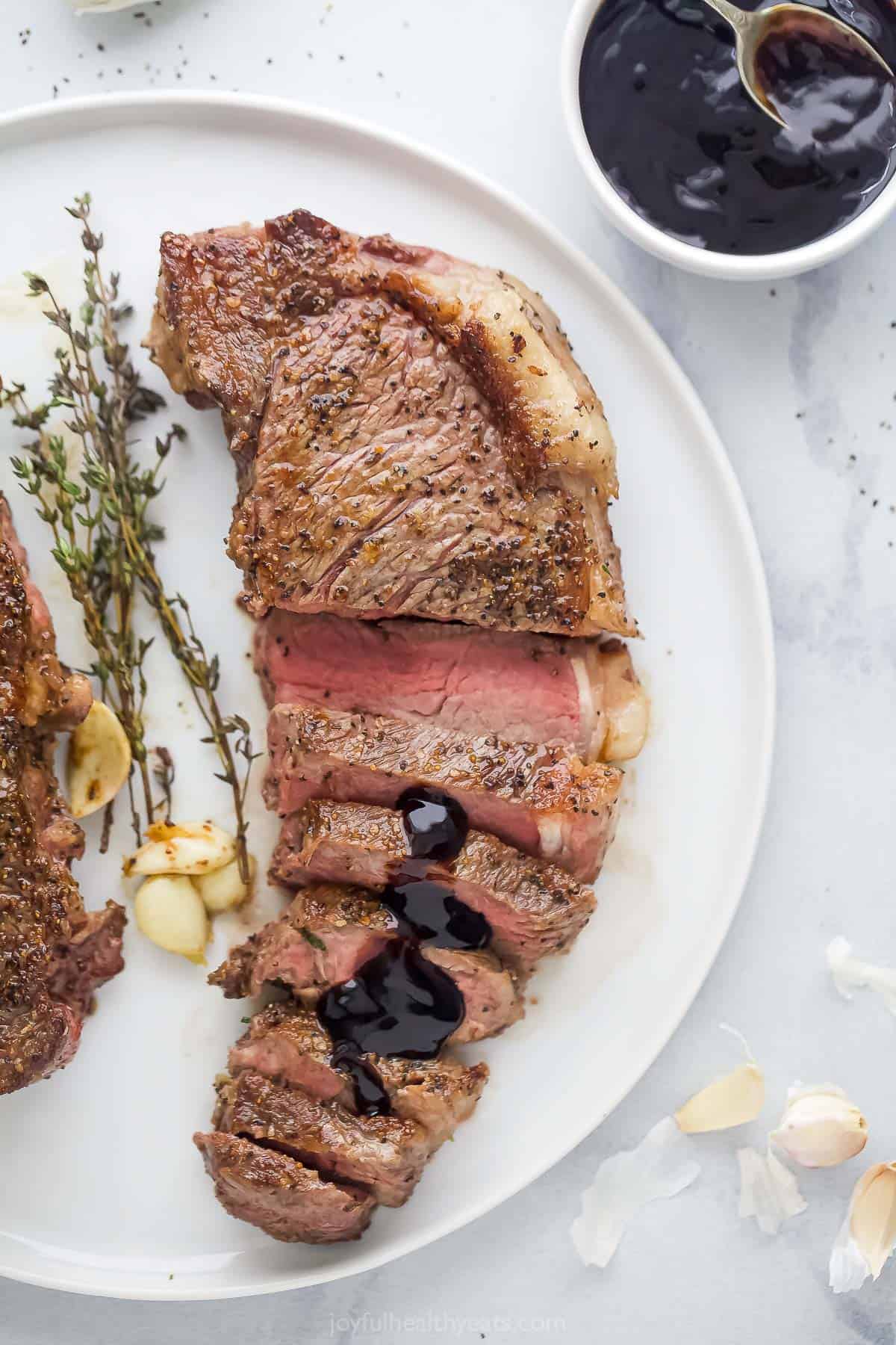 This pan-seared New York strip steak recipe with garlic-herb butter and balsamic reduction is an easy, fancy dinner you'll love. Sliced New York strip steak served on plate with balsamic reduction drizzled on top.