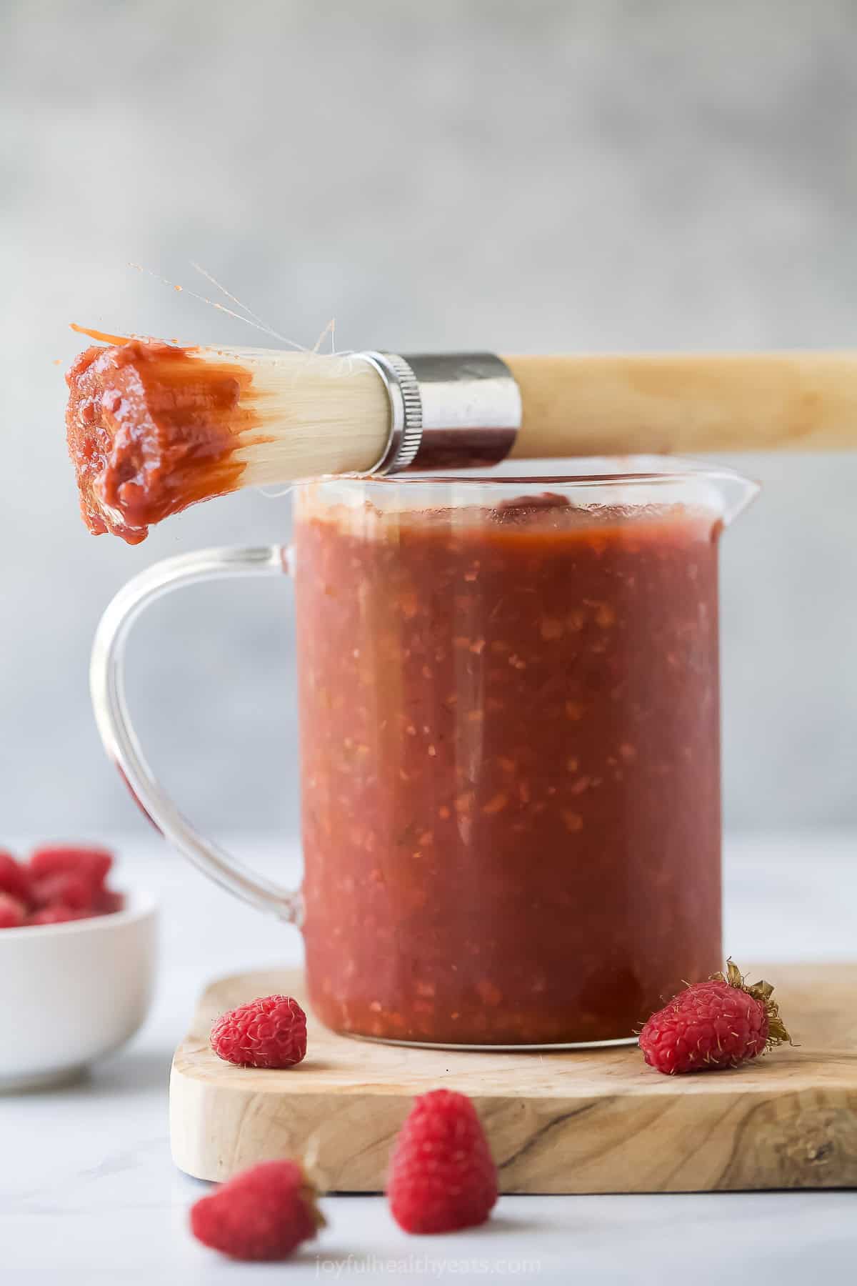 This smokey raspberry chipotle BBQ sauce recipe with garlic, liquid smoke, and sweet onion goes great with your favorite cookout dishes. Small pitcher of raspberry chipotle BBQ sauce with a brush on top.