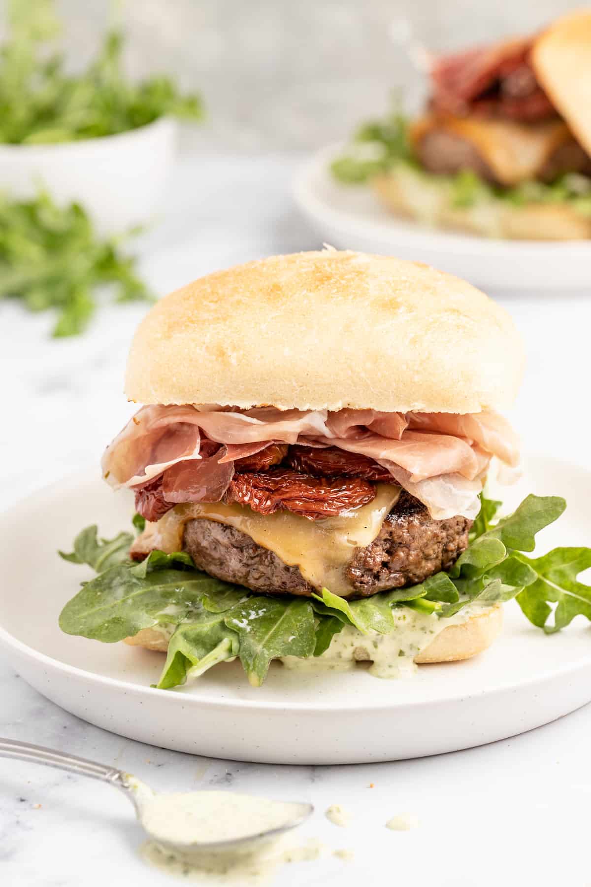 This 15-minute Italian cheeseburger is loaded with gouda cheese, sun-dried tomatoes, pesto aioli, and prosciutto for the perfect bite. Italian cheeseburger on a plate.