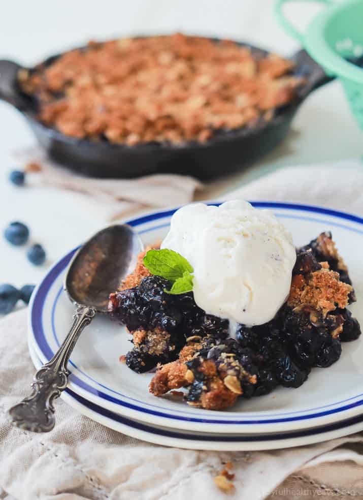 Juicy blueberries combine with sweet ginger and a buttery cinnamon pecan topping in this easy blueberry crisp! It's a summertime classic. A serving of berry crisp on a plate with the remaining crisp inside of a pan in the background.