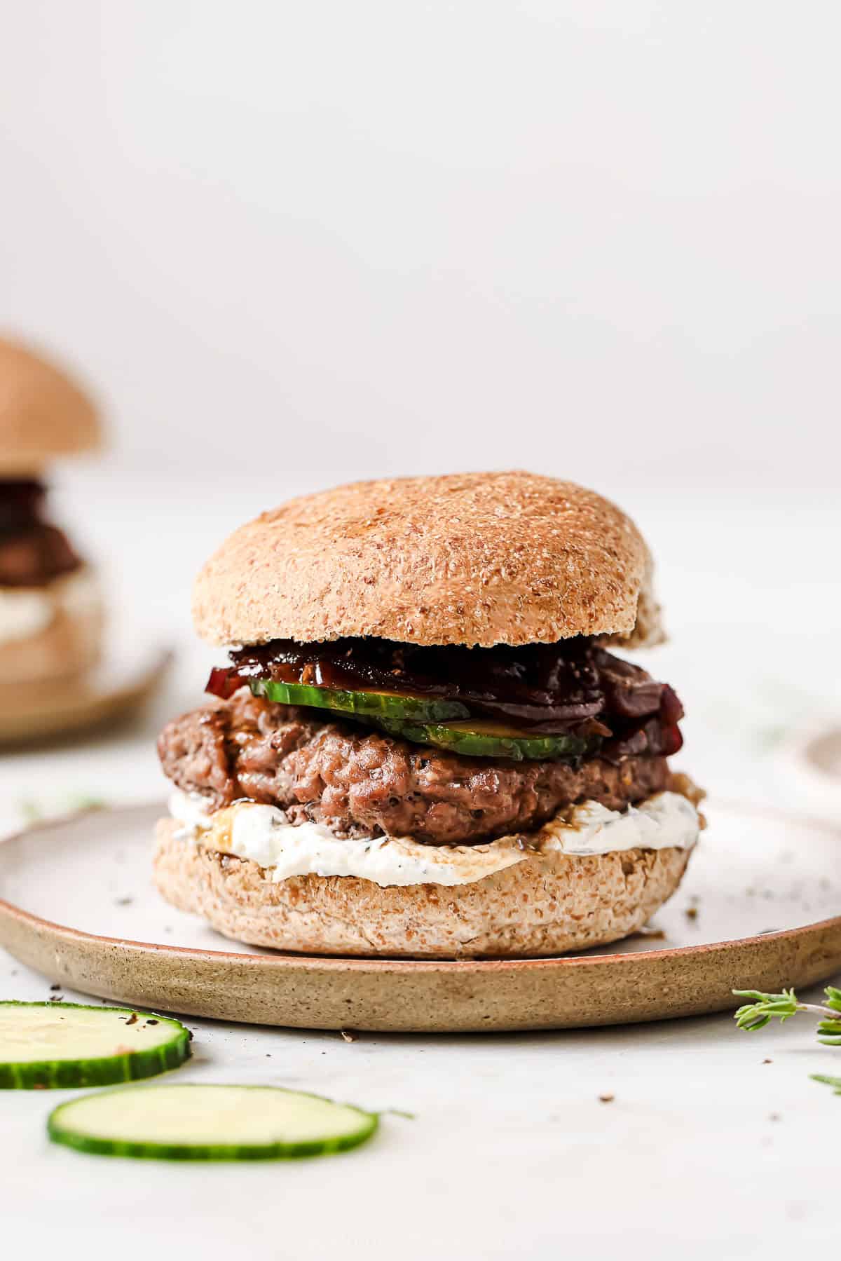A lamb burger with whipped feta, caramelized onions, and sliced cucumbers on a serving plate. 