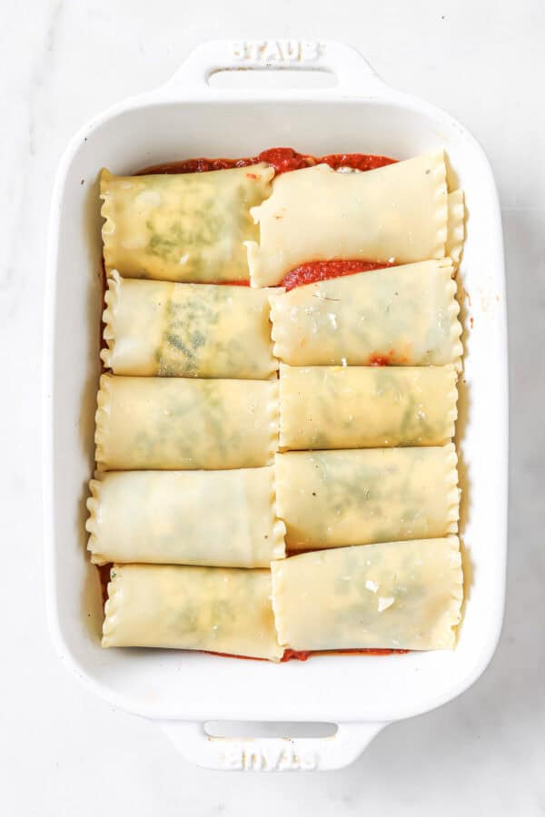 Rolled lasagna sheets with cheese filling in the baking tray with a layer of marinara sauce on the bottom. 