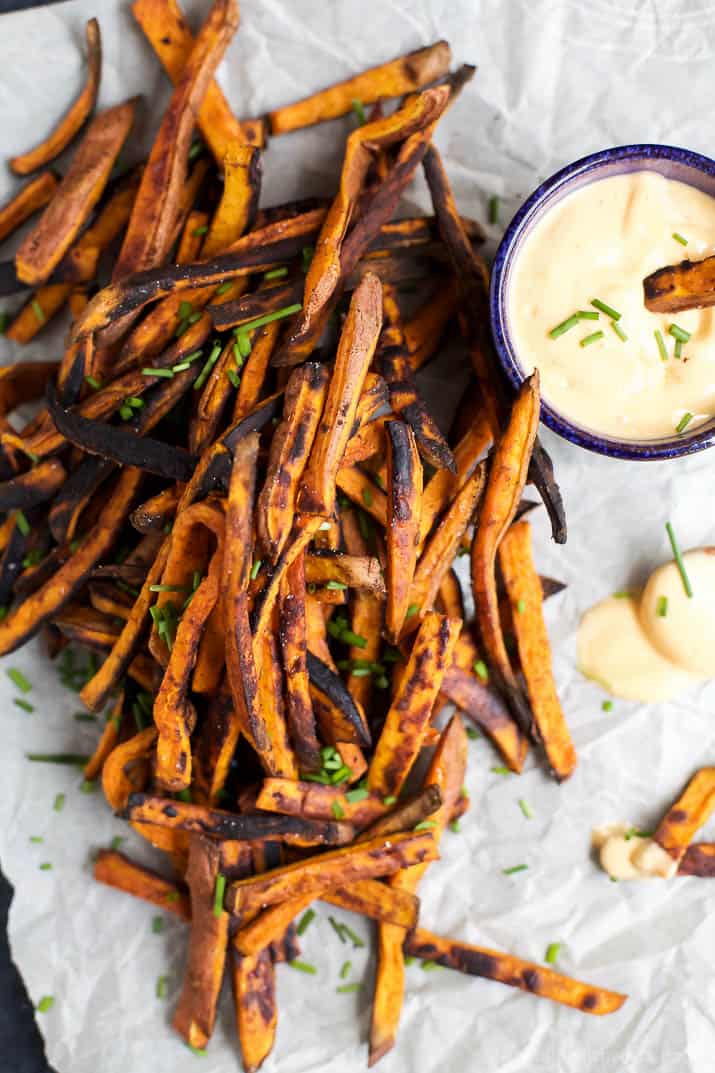 An overhead view of a batch of sweet potato fries on a sheet of parchment paper with honey sriracha sauce beside them.