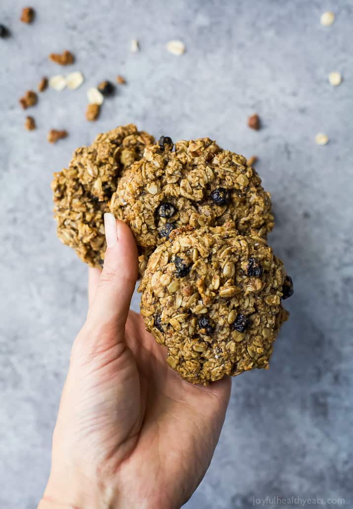 These moist & chewy Blueberry Breakfast Cookies feature heart-healthy oats, high-protein almond butter, antioxidant-rich chia seeds and more! A hand holding three blueberry oatmeal cookies above a kitchen countertop