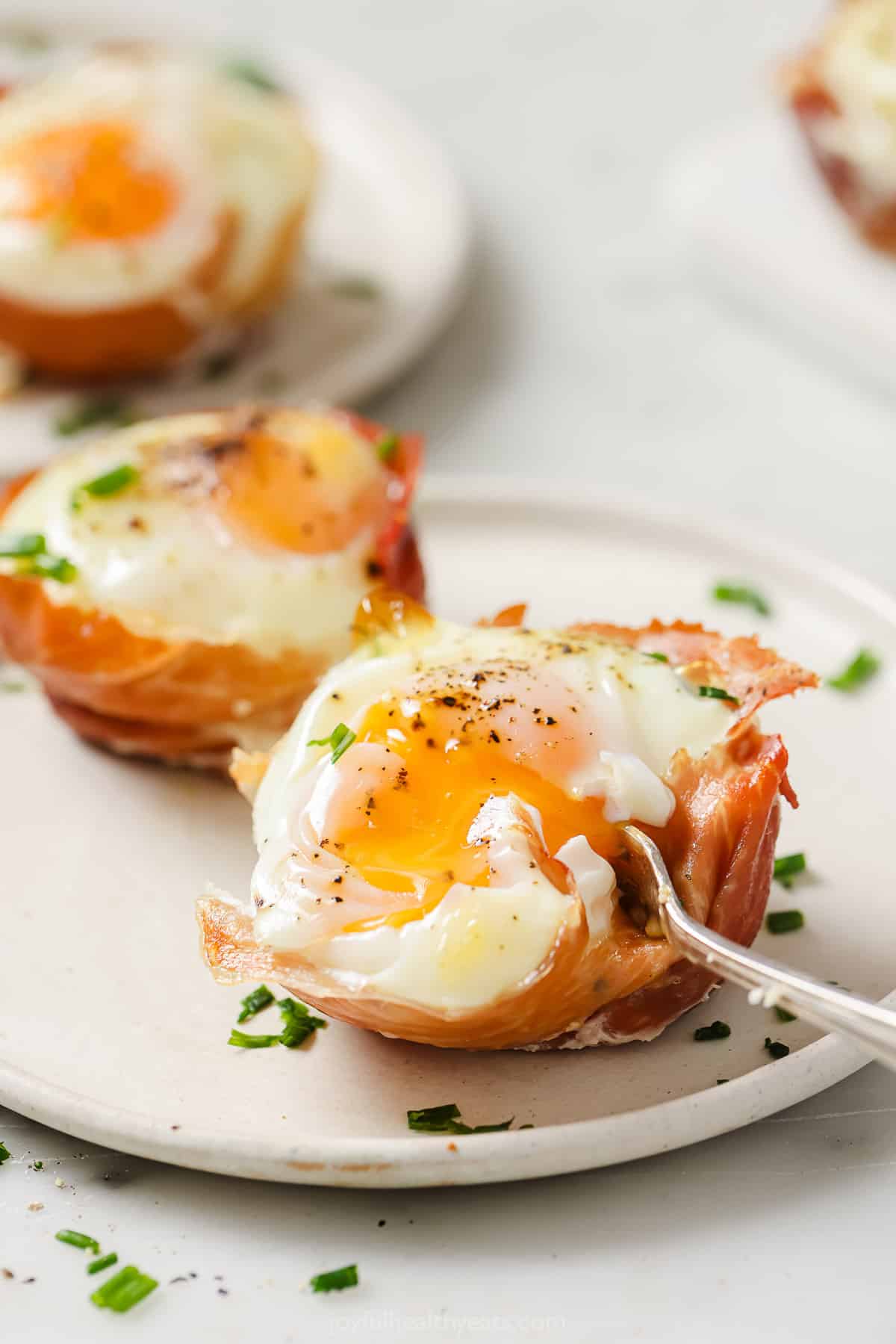 This baked egg cups recipe with crispy prosciutto shells, garlicky spinach, and tender bell peppers is the perfect easy breakfast. Digging into a prosciutto egg cup with a fork, revealing a runny yolk.