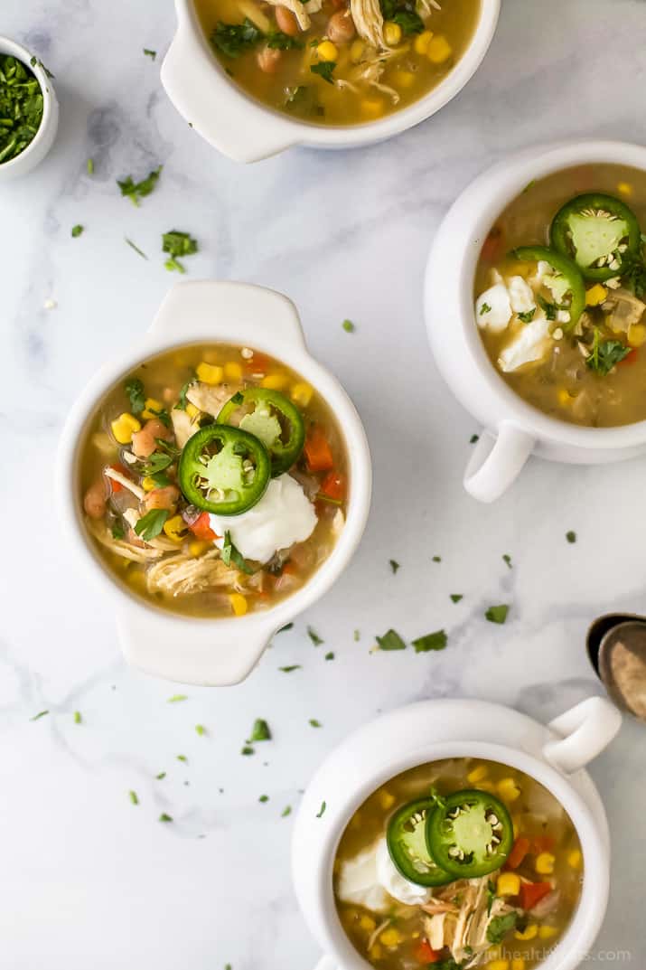 This healthy white chicken chili recipe is filled with juicy chicken, hearty pinto beans, tender corn, and more. Perfect for chilly days! Top view of crocks of white chicken chili with jalapeno slices and sour cream.