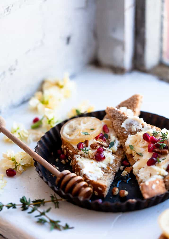 A serving plate with two halves of ricotta toast with pomegranate, honey, candied pecans, and fresh thyme. 