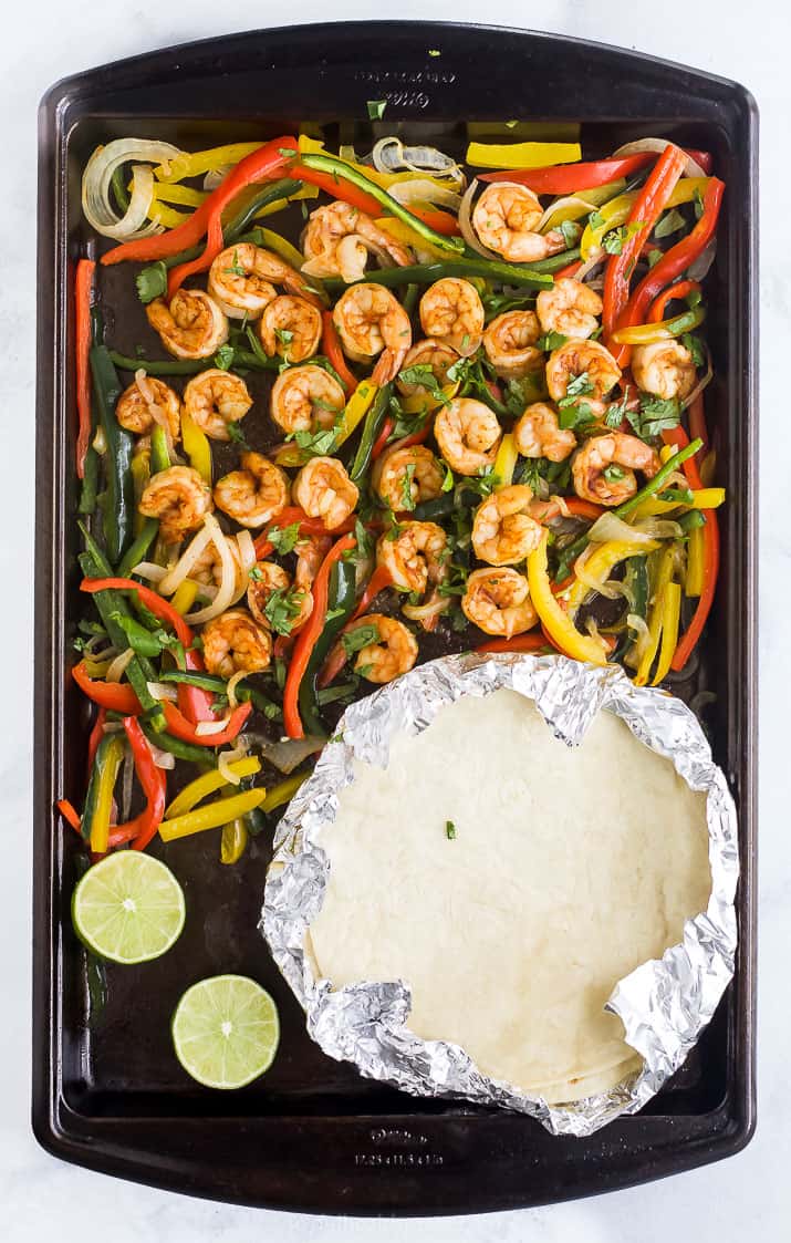 Tortillas wrapped in foil on a baking sheet with homemade baked shrimp fajitas. 