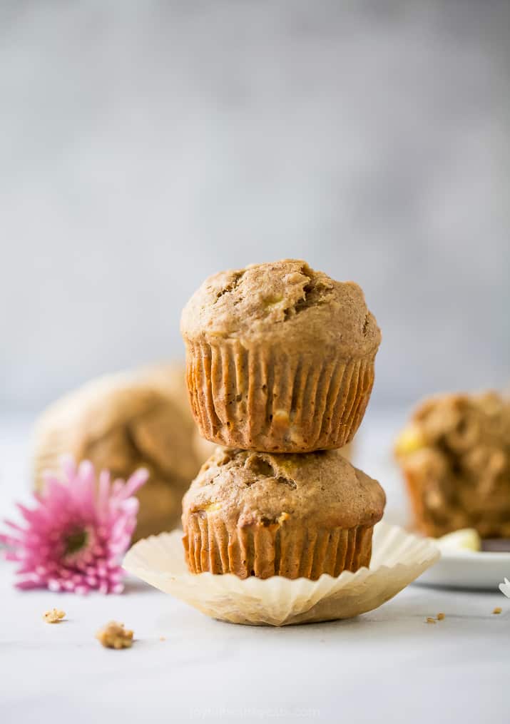 These whole wheat banana muffins are loaded with banana flavor, cinnamon and nutmeg! They're moist, fluffy & perfect for making ahead. A stack of two whole wheat banana muffins on a countertop with a pink flower beside them