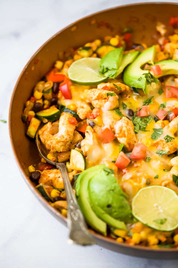 Close up of a chicken enchilada skillet topped with melted cheese, garnished with avocado slices and lime.
