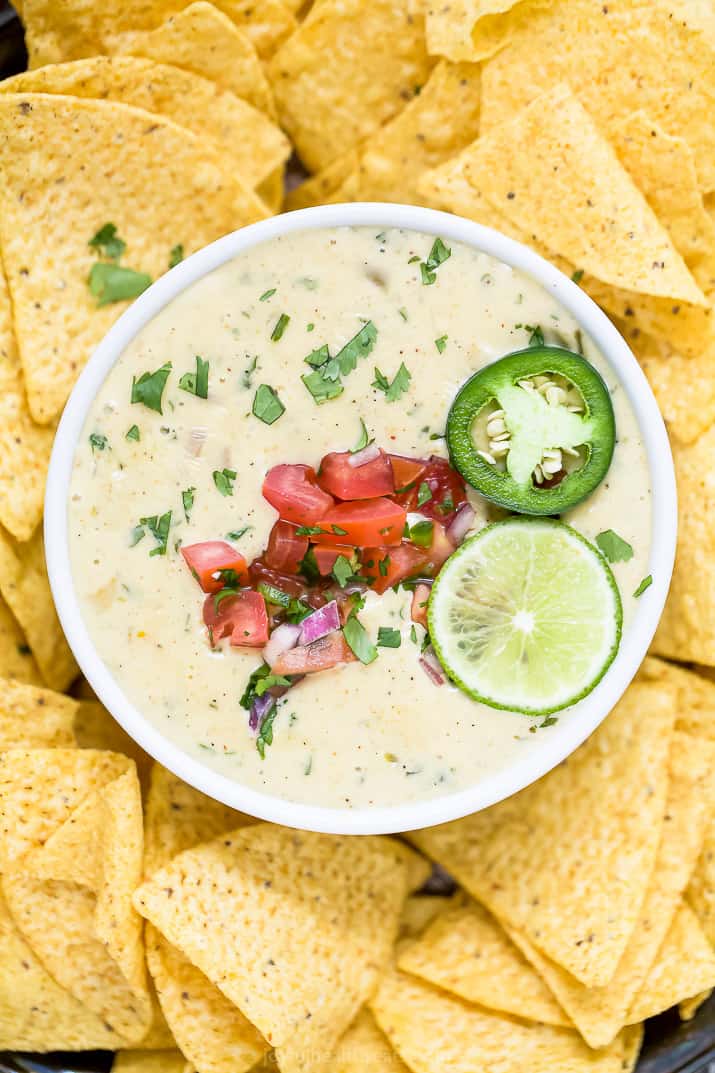 This easy white queso dip recipe is a classic Mexican appetizer made from melty white cheese and chilies, garnished with pico de gallo. Overhead view of a bowl of white queso dip garnished with lime and pico de gallo surrounded by tortilla chips.
