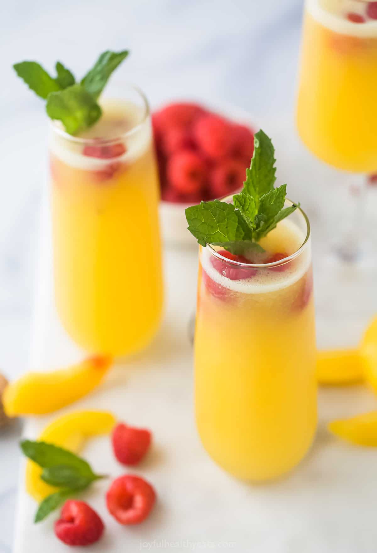 Two peach cocktails on a marble surface with peach slices, raspberries and mint sprigs