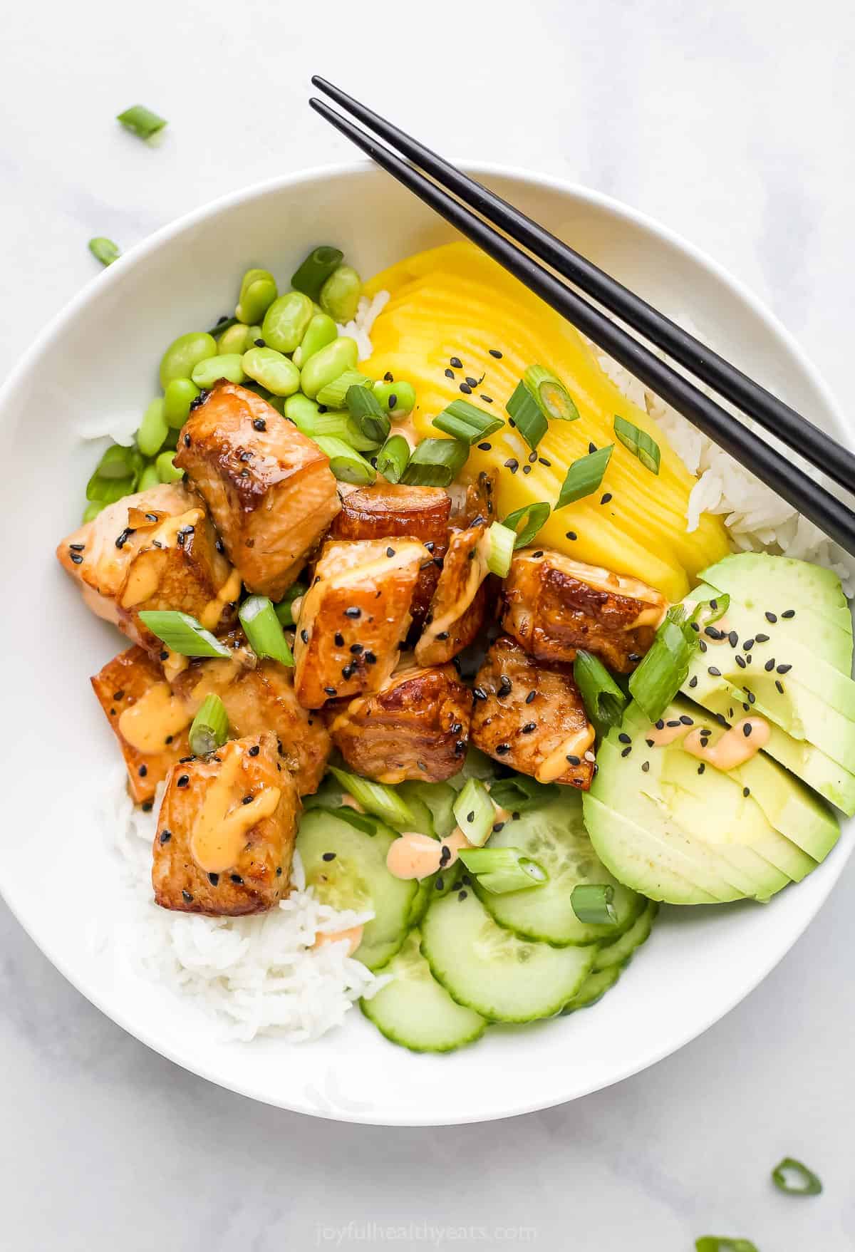 This Salmon Poke Bowl recipe is made with marinated baked salmon, cucumbers, mango, edamame & avocado on a bed of basmati with spicy mayo. A close-up shot of a rice bowl with fish, mango, avocado, cucumber and more