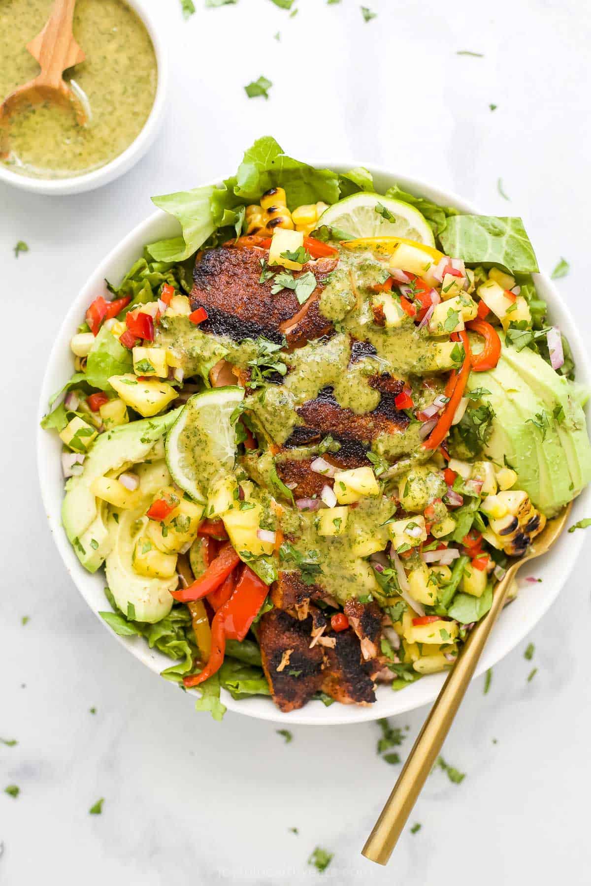 Smoky blackened salmon mixes with crunchy lettuce, juicy corn, creamy avocado, fresh pineapple salsa, and more in this grilled salmon salad! A bowl of salmon salad on a kitchen countertop beside a small dish of cilantro lime dressing.