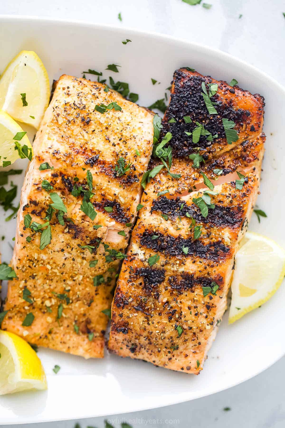 Tangy lemon pepper seasoning makes tender grilled salmon taste better than ever! Plus, each fillet develops just the right amount of char. Zesty grilled lemon pepper salmon is a must-make recipe! Two grilled citrus salmon fillets on a plate on top of a marble counter.