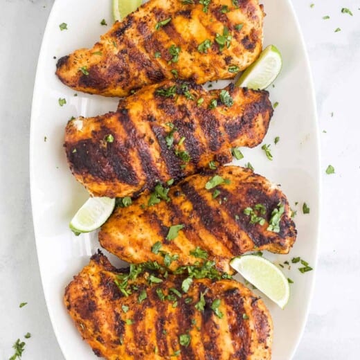 Four grilled chicken breasts on a serving platter with a kitchen towel and a dish of salt beside it