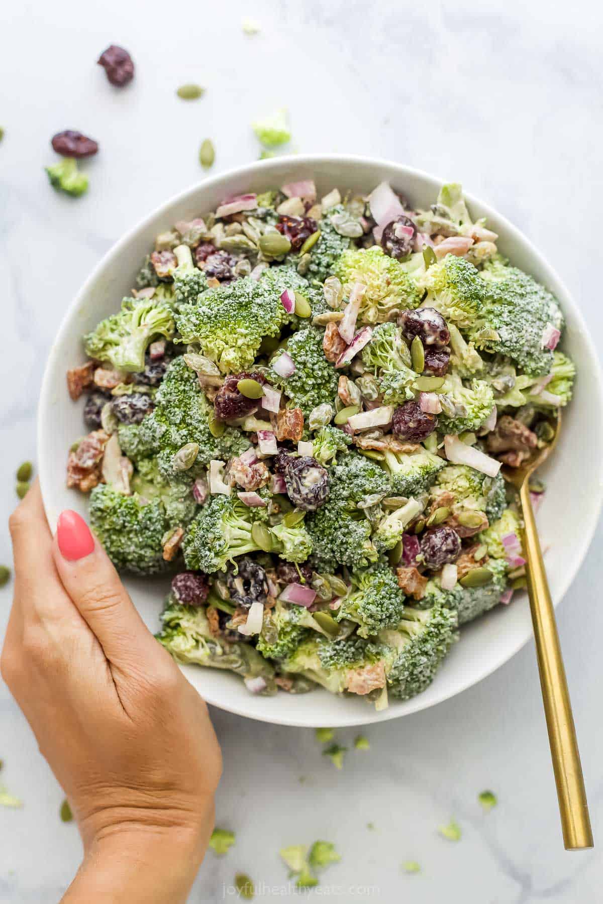 Lightened-Up Broccoli Salad With Bacon | Joyful Healthy Eats A hand holding a big bowl of broccoli salad with a gold-colored spoon inside