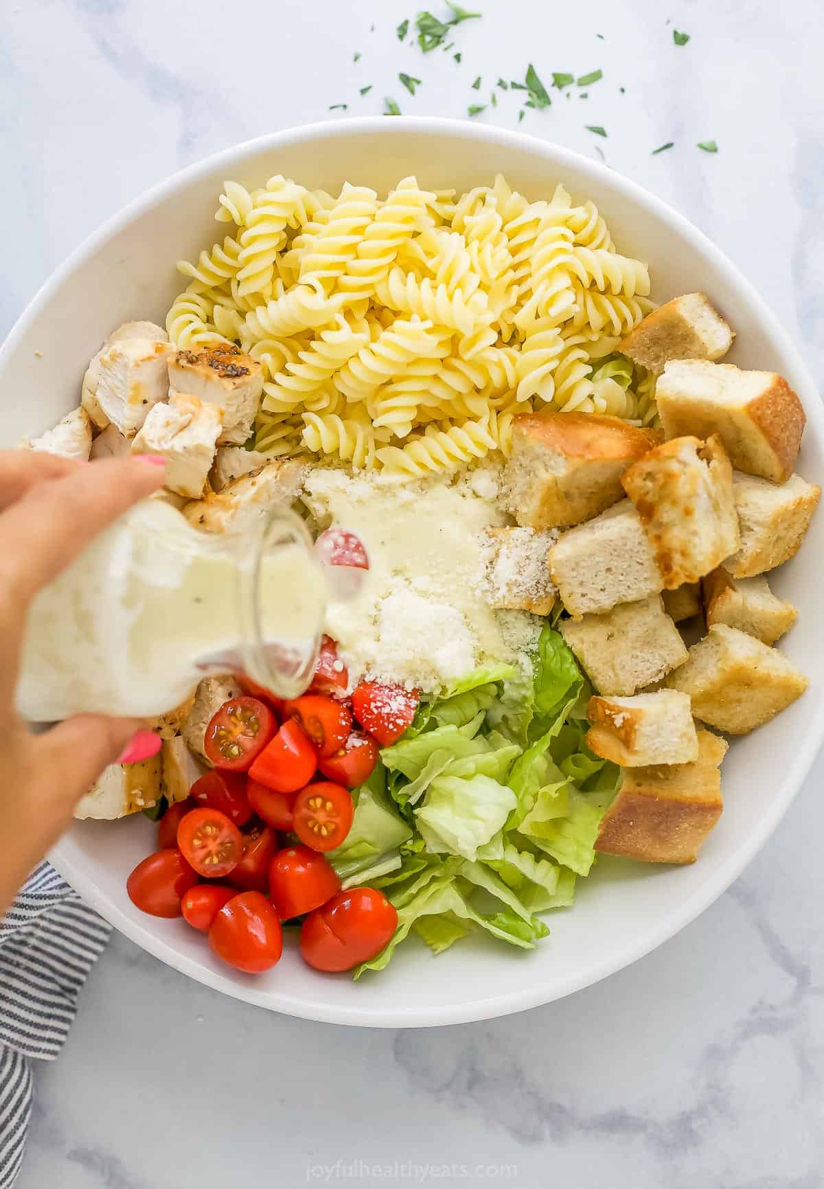 Caesar dressing being poured over a bowl full of rotini noodles, homemade croutons and the rest of the salad ingredients