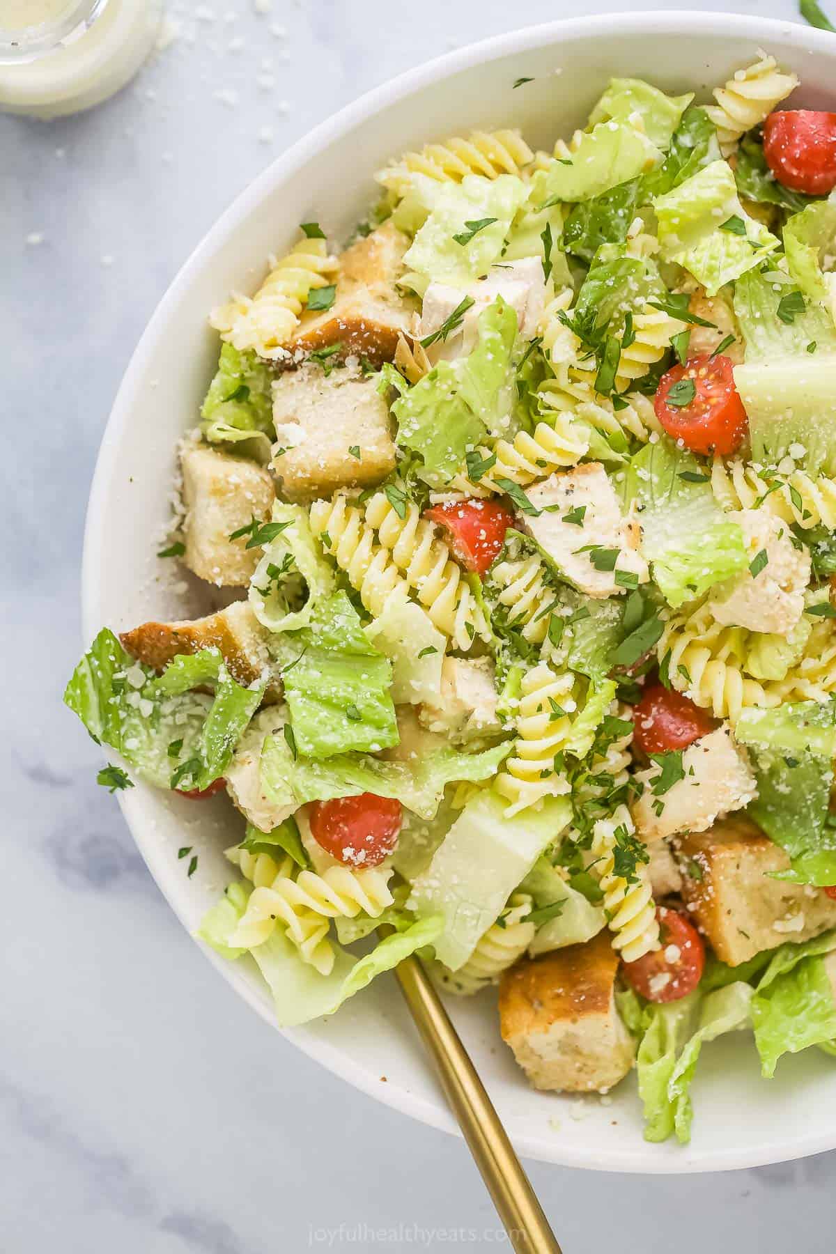 Tender chicken, fresh lettuce and chewy noodles combine with crunchy croutons and creamy Caesar dressing in this hearty Chicken Pasta Salad! A chicken caesar pasta salad in a big white bowl with a bottle of homemade caesar dressing beside it