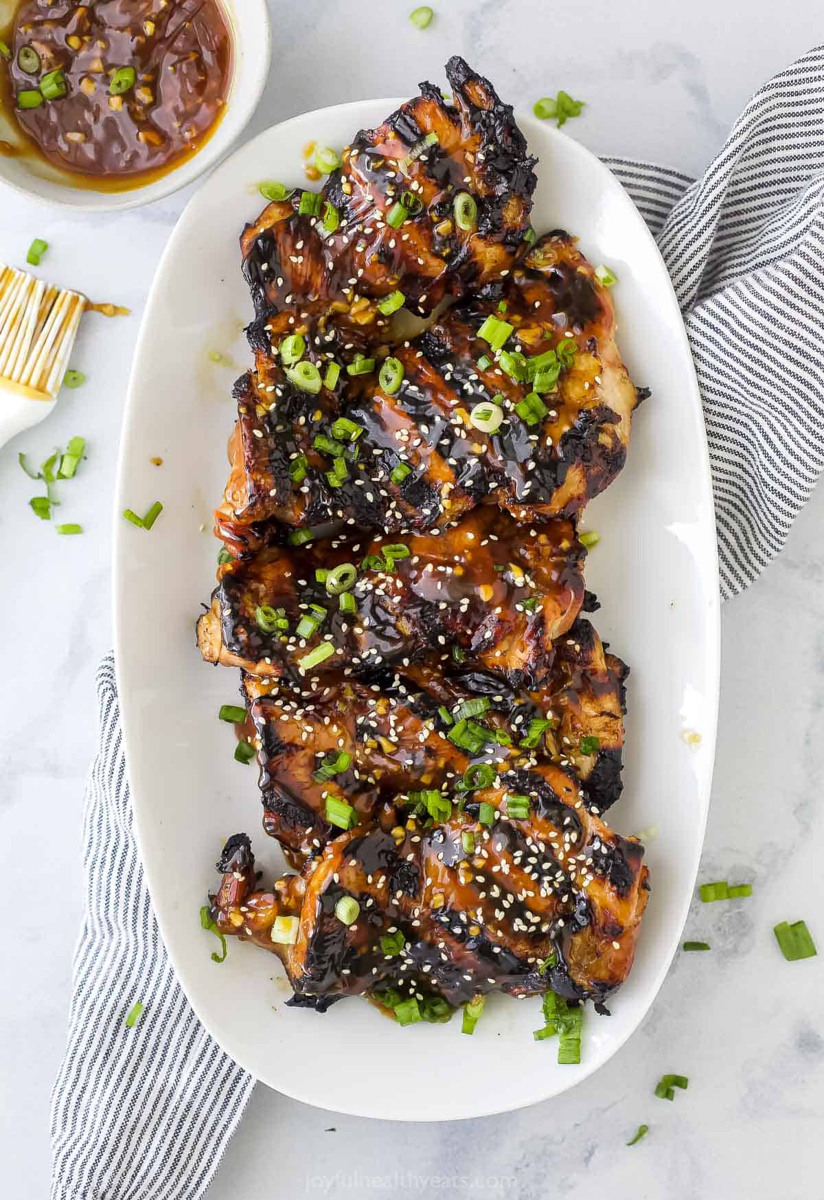 Tender chicken thighs are soaked in a flavorful honey teriyaki marinade and seared to perfection over the grill in this foolproof recipe. A platter full of teriyaki grilled chicken thighs on top of a striped kitchen towel