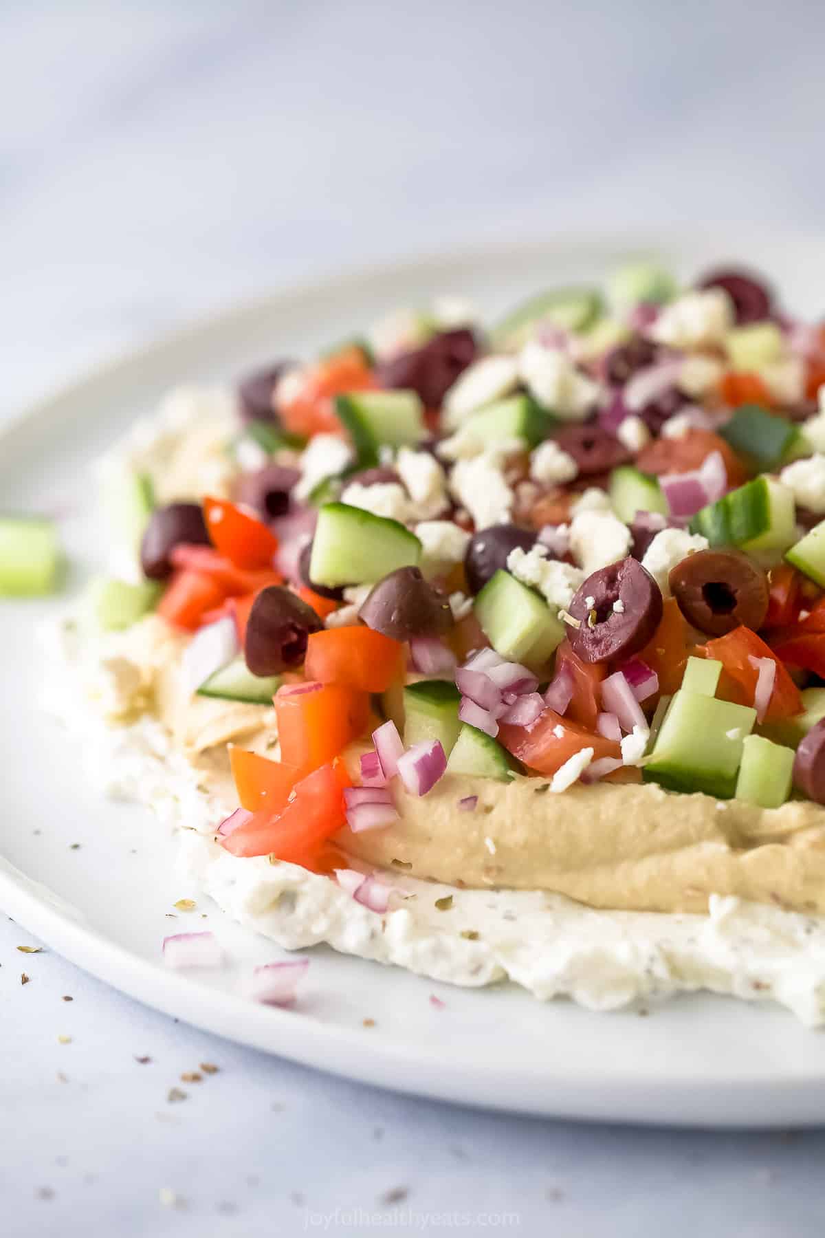 Colorful veggies combine with smooth hummus and fluffy whipped feta cheese in this classic Mediterranean 7 Layer Dip. It's made in 5 minutes! A close-up shot of Mediterranean 7 layer dip on a flat white plate