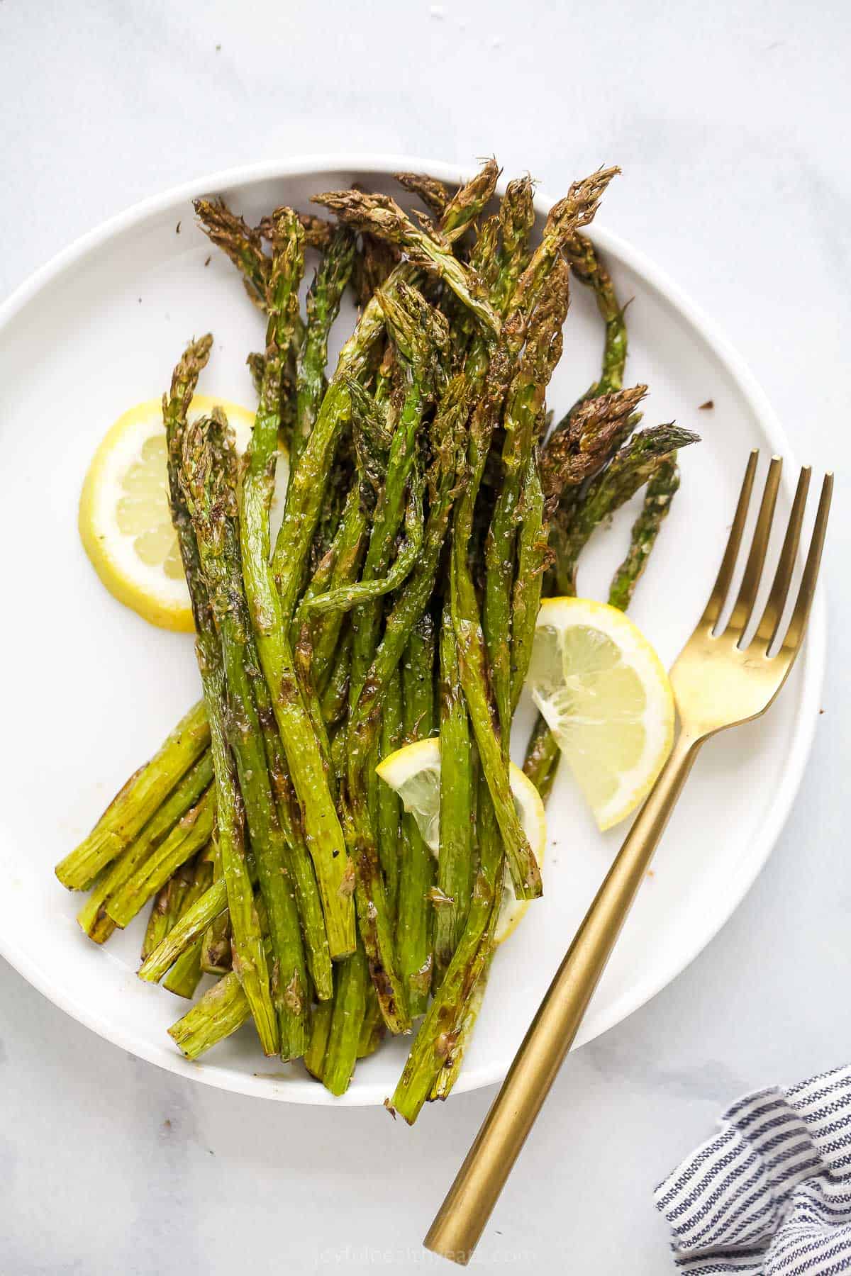 Gently seasoned with salt, pepper and garlic powder, this tender-crisp Air Fryer Asparagus doesn't need anything fancy to make it shine! Air-fried asparagus on a white plate with fresh lemon slices and a golden fork