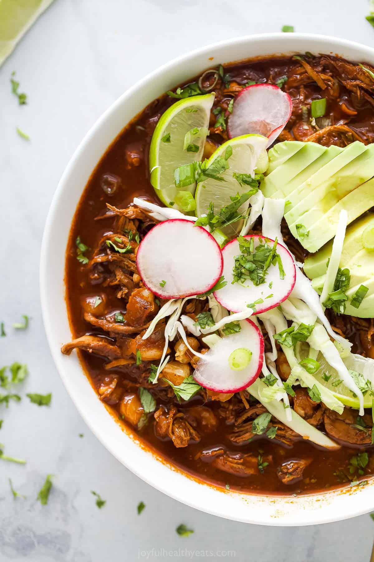Chicken Pozole Rojo is a Mexican stew with chicken, hominy, and red enchilada sauce. The stew is simple and has incredible authentic flavors. a bowl of red sauce based chicken stew garnished with radishes, lime wedges, cilantro, and avocado slices