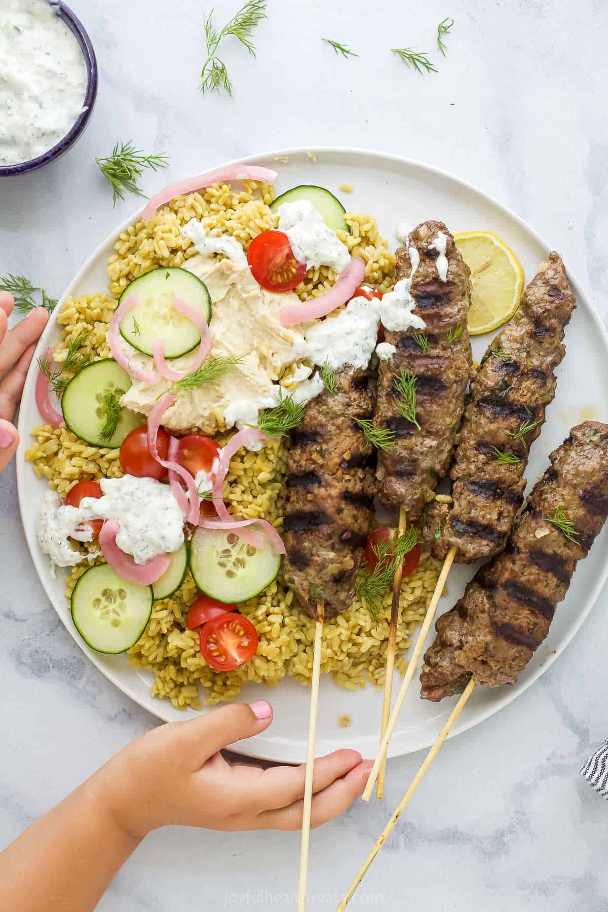 Ground beef is blended with diced onions, minced garlic and a flavorful Middle Eastern spice mix in these tender pan-seared Kefta Kabobs. Four seared beef kabobs over rice on a plate with veggies and tzatziki sauce