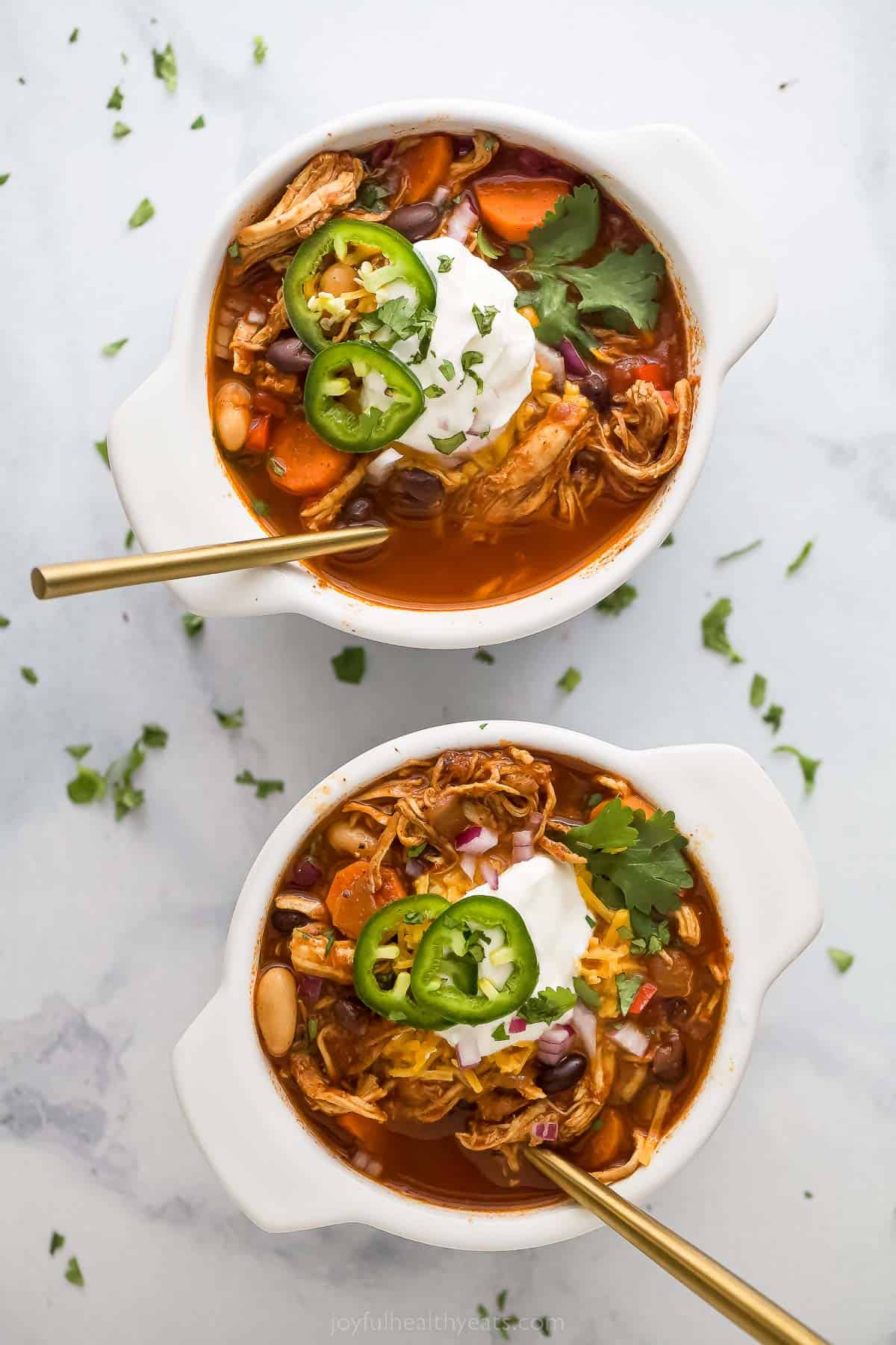 Smoky chicken chili is the ultimate comfort food, loaded with fresh vegetables, hearty beans, chicken, and rich, smoky chipotle peppers. Two bowls of chicken chili with jalapeno, sour cream, and cilantro garnish.