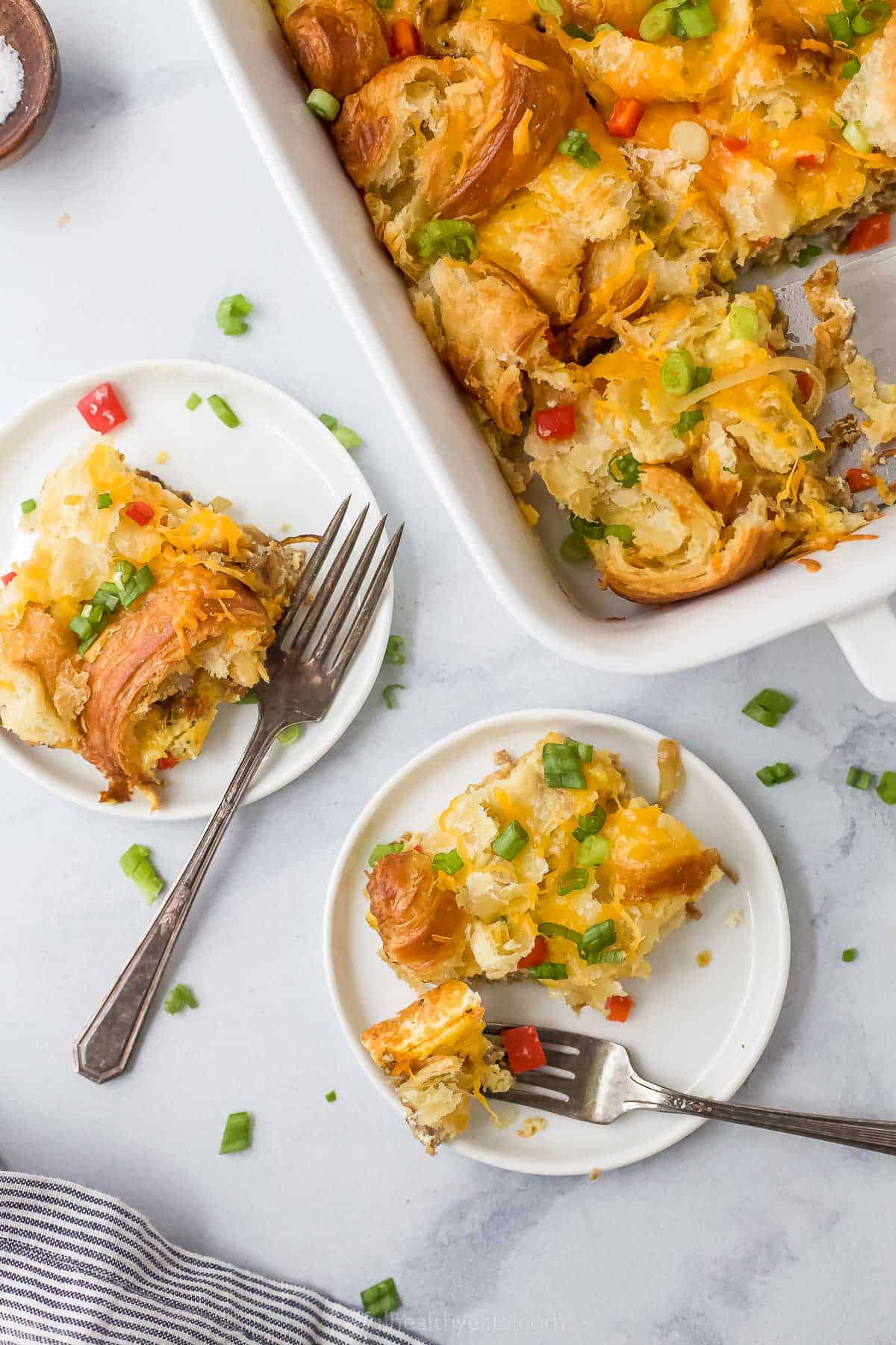 This easy Croissant Breakfast Casserole has all the breakfast flavors you love. It has whisked eggs, sausage, cheese, onions, and peppers. two plates next to a casserole dish with croissant breakast bake with sausage
