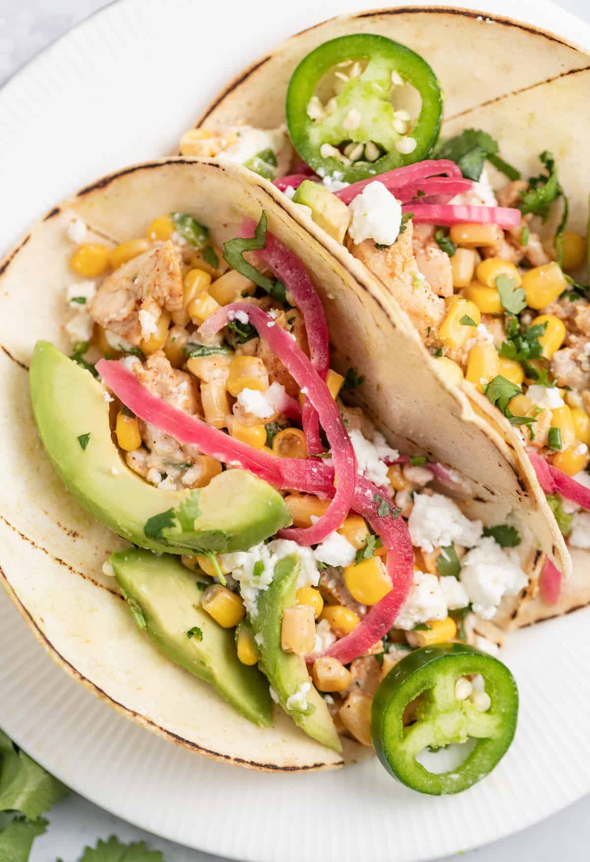 These Chicken Street Tacos are topped with Mexican Street Corn, avocado, and Homemade Pickled Onions. The perfect recipe for taco night. two plated Chicken Street Tacos with avocado, pickled onions, and corn salad