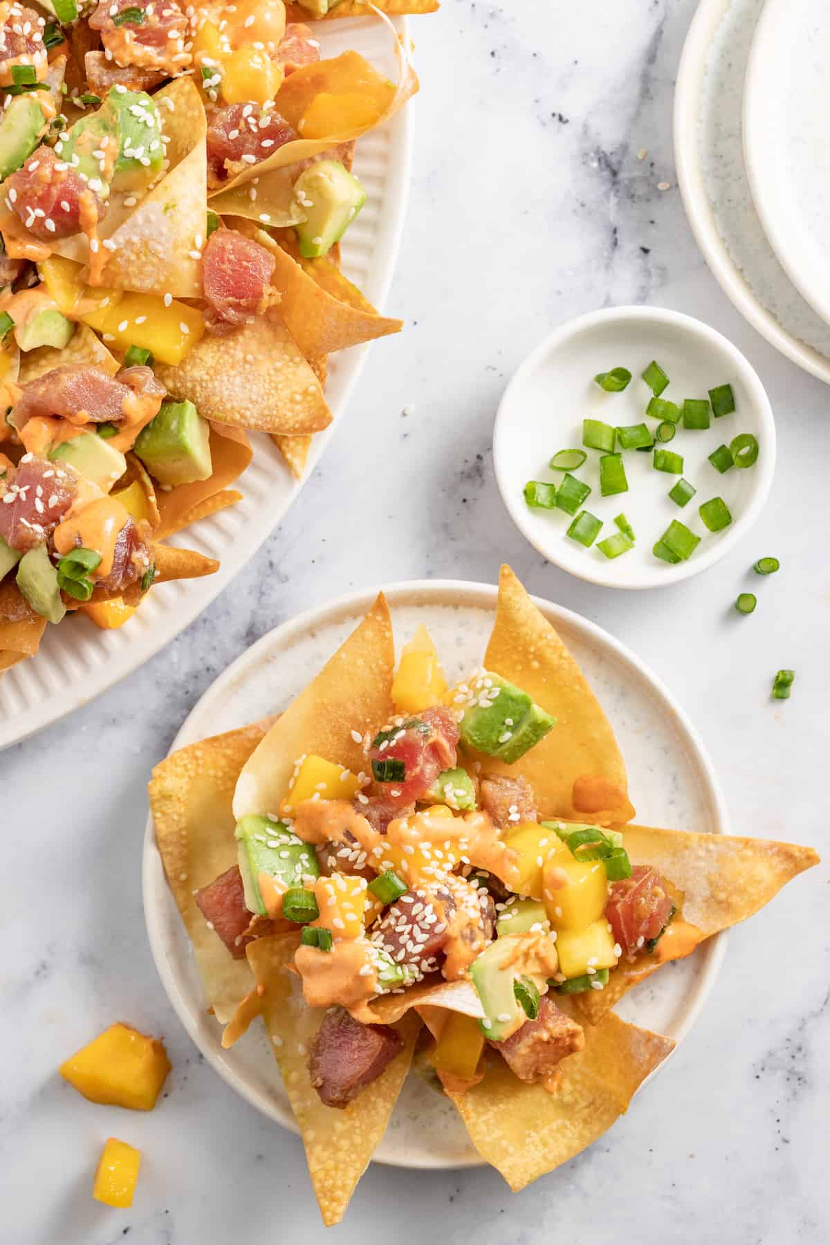 Ahi Tuna Poke Nachos are a unique shareable snack. They are made with homemade wonton chips, seasoned ahi tuna, mango, and avocado. a plate of ahi tuna poke nachos with diced mango, green onions, and sriracha mayo