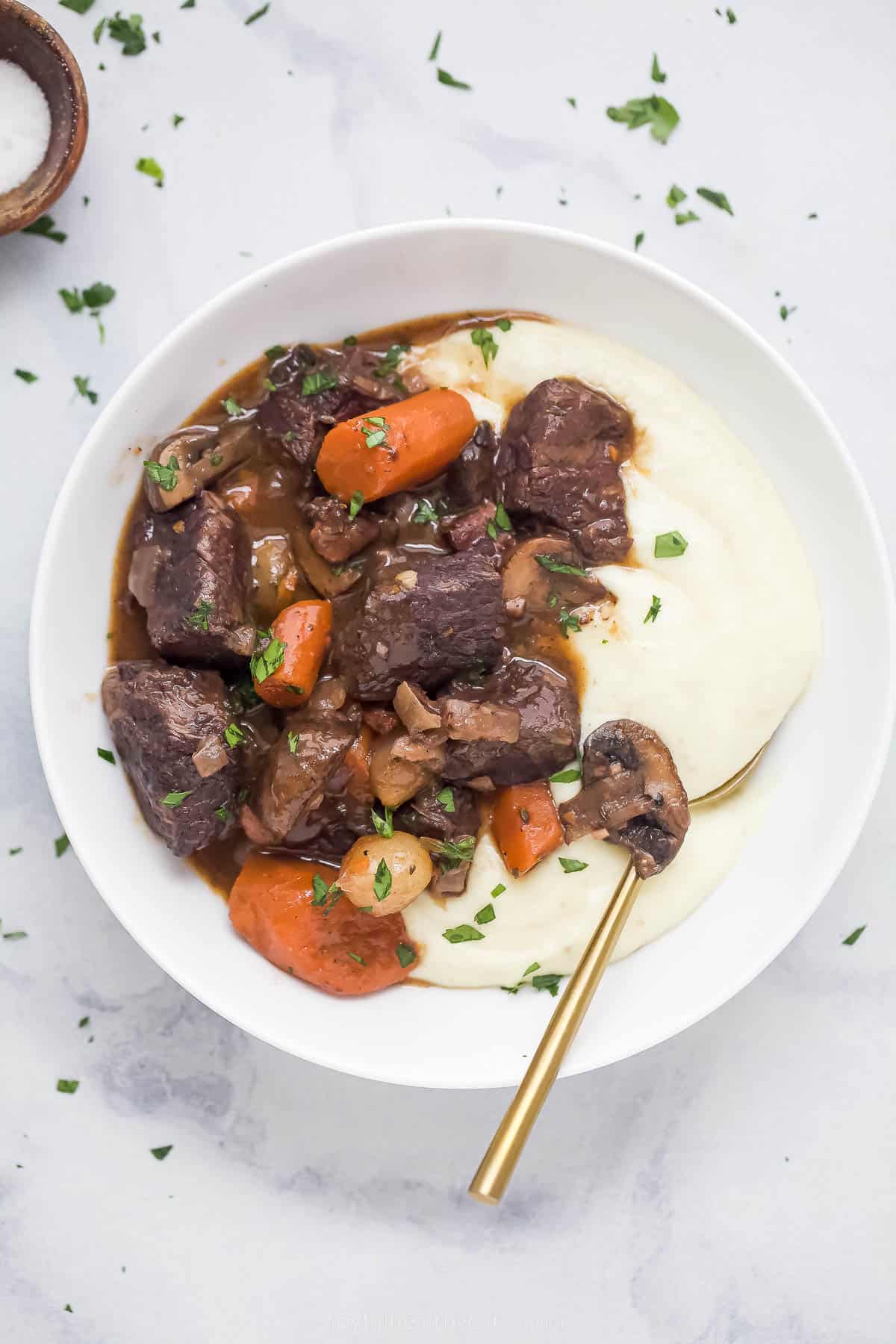 This Beef Bourguignon recipe is a classic French dish that features tender beef, cooked vegetables, and a rich and herbal gravy. chunks of cooked beef with cooked carrots and onions on top of pureed potatoes