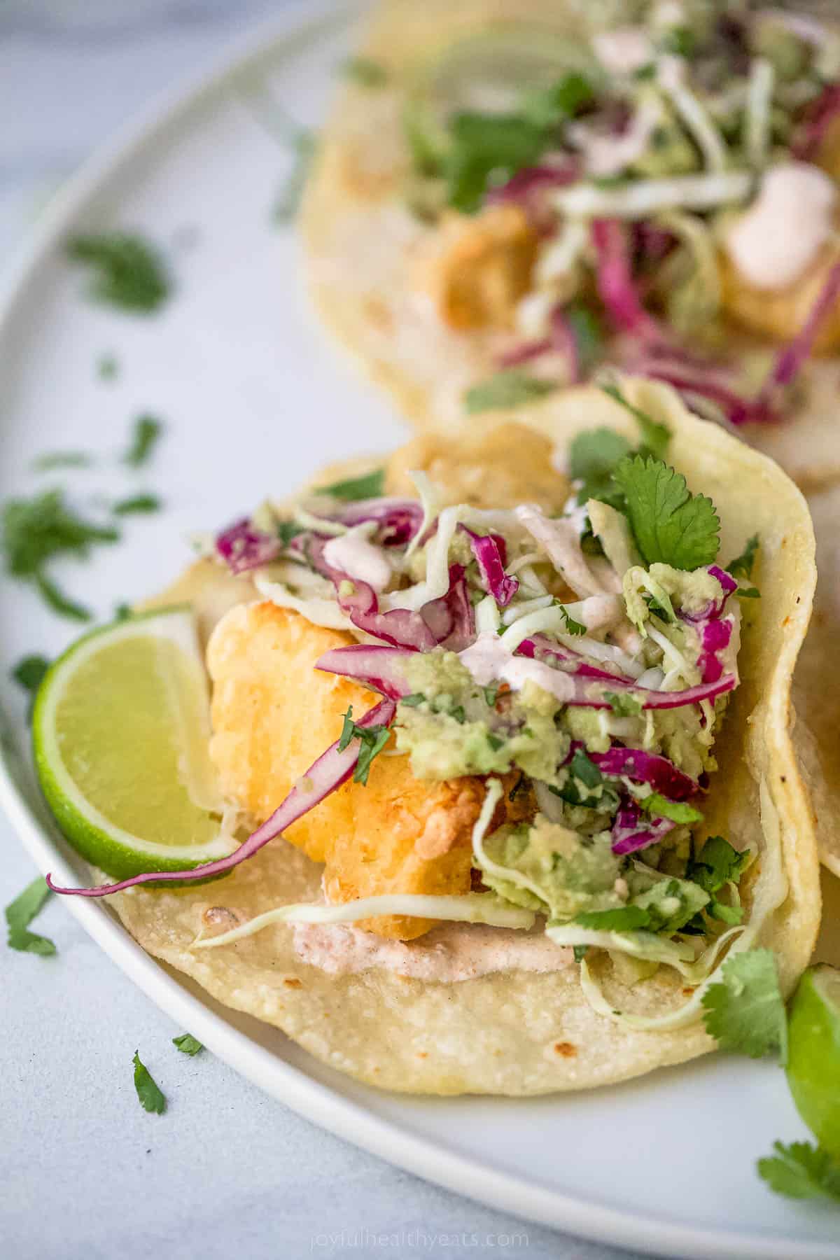 Beer Battered Air Fryer Fish Tacos are dipped in a tasty batter and fried the healthy way! Add slaw and Chipotle Crema for the perfect taco! a fish taco on a plate with lime garnish and slaw on top