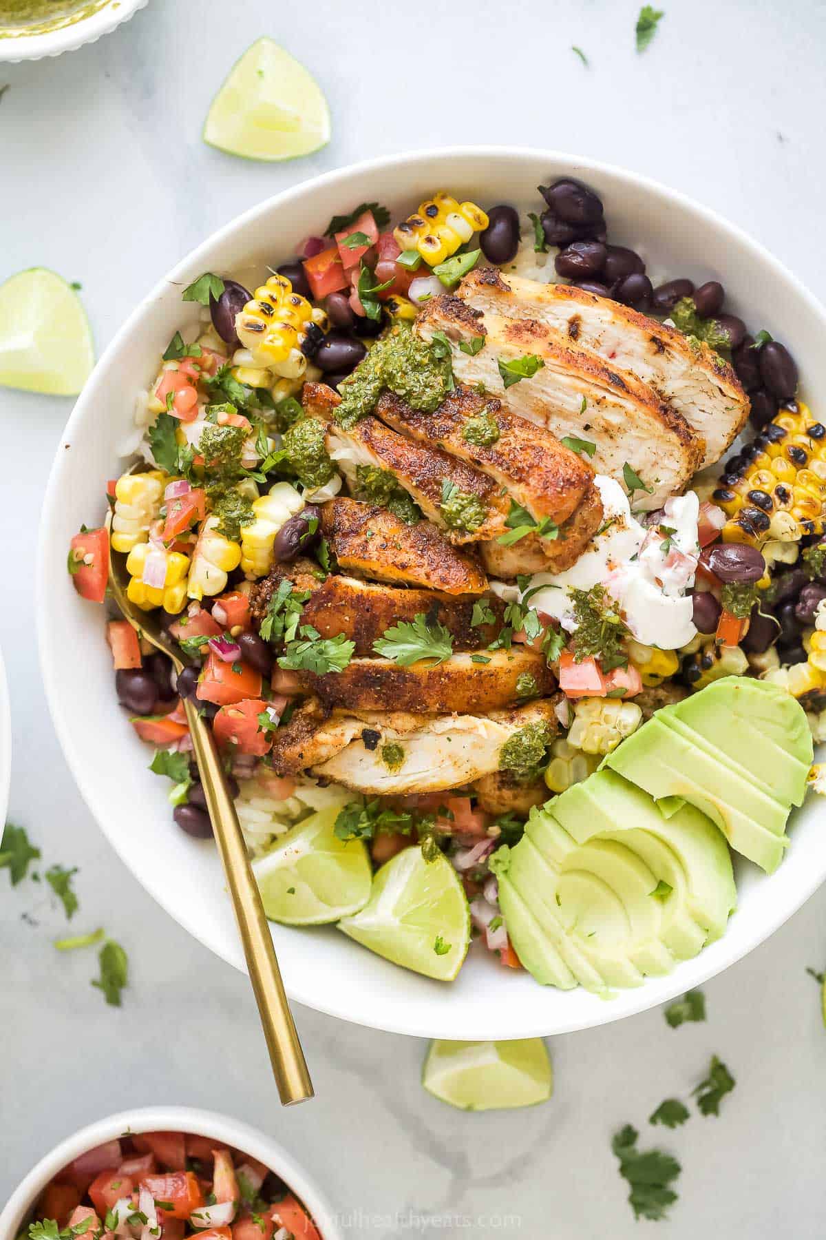 This easy chicken burrito bowl recipe is a hearty Tex-Mex dinner packed with juicy chicken, fresh veggies, and homemade chimichurri. Chicken burrito bowl with avocado, lime wedges, and other toppings.