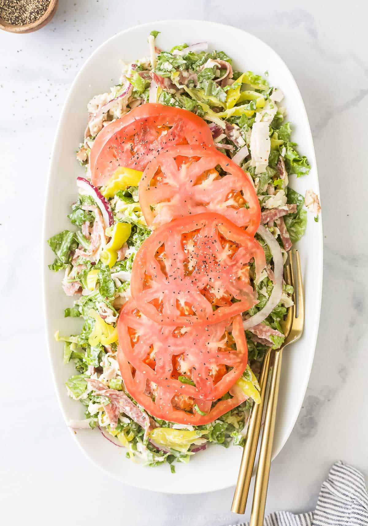 A take on the viral grinder sandwich, this Italian grinder salad is made with sliced cold-cuts, provolone, onion, pepperoncini, tomatoes, and a grinder dressing! It's the ultimate 15-minute salad. Plate of Italian grinder salad with large tomato slices on top.
