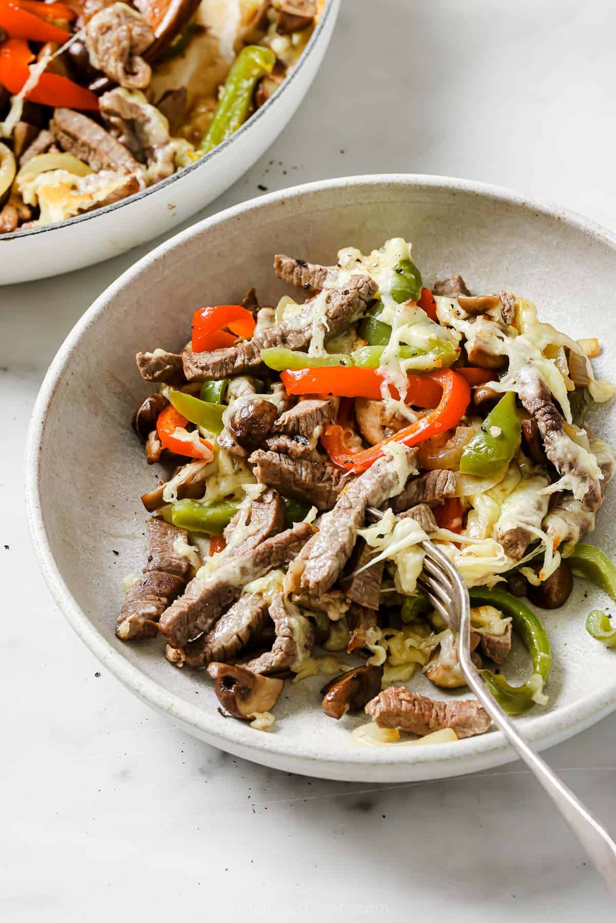 This one-pan philly cheesesteak recipe is packed with garlic, tender peppers, juicy steak, and mushrooms for the perfect weeknight dinner. Philly cheesesteak skillet in a bowl with melty mozzarella on top.