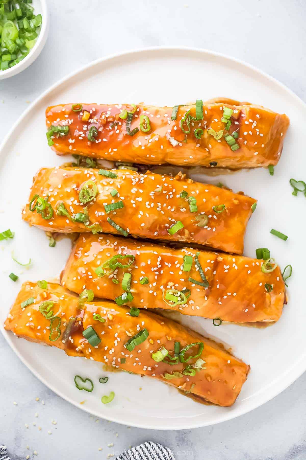 This saucy teriyaki salmon recipe with crunchy green onions and fluffy white rice is the perfect take-out style dinner. Teriyaki salmon with green onions and sesame seeds on a plate.