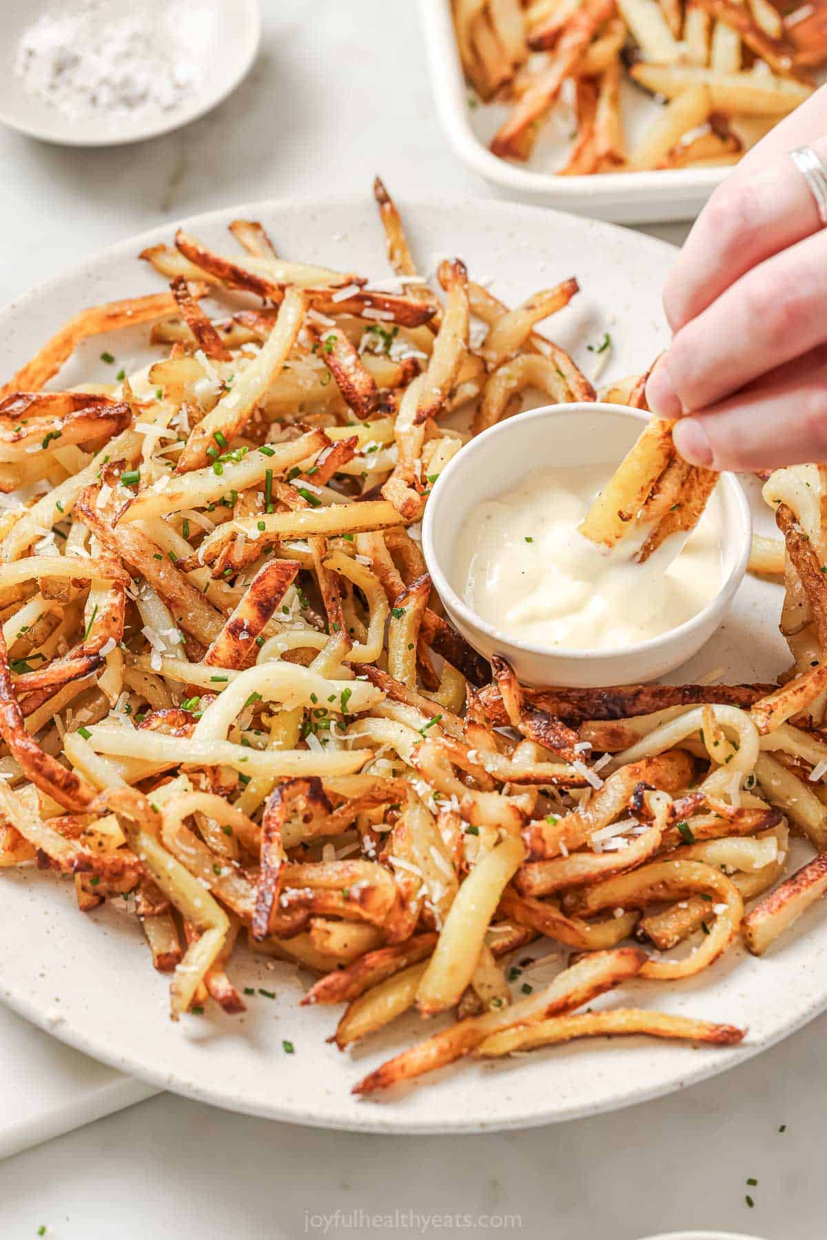 Overhead view of baked Parmesan truffle fries on platter, with hand dipping fries into aioli